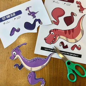 Puede incluir: Un proyecto de manualidades para niños con recortes de dinosaurios. La imagen muestra hojas impresas con partes de dinosaurios, incluyendo un Parasaurolophus morado y un T-Rex rojo. También hay unas tijeras con mangos verdes, junto con el texto "CUT AND GLUE".