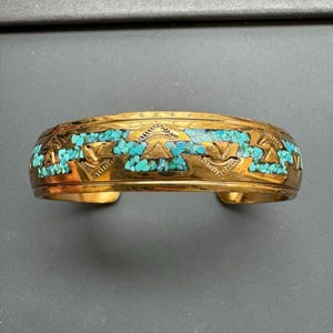 May include: A gold-toned cuff bracelet with turquoise inlay. The bracelet features a geometric design with turquoise stones set between decorative elements. The open cuff design allows for easy wear. The bracelet is a statement piece.