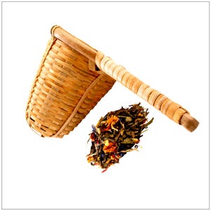 May include: A woven bamboo tea strainer with a long handle, next to a pile of loose leaf tea with colorful flower petals. The strainer is light brown and appears handmade.