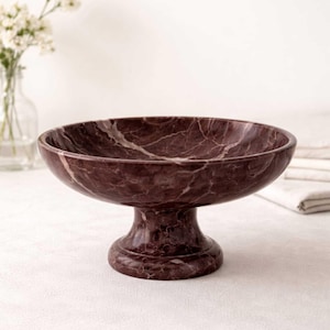 May include: A burgundy marble pedestal bowl with white veining. The bowl has a wide, shallow basin and a rounded base. The bowl is displayed on a white surface, with a vase of flowers in the background.