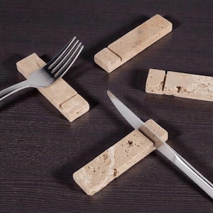 Puede incluir: Descansos para tenedores y cuchillos de piedra beige. Los descansos rectangulares tienen una ranura para sujetar la cubertería. Un tenedor y un cuchillo de acero inoxidable se colocan en dos de los descansos. Los descansos están sobre una superficie marrón oscuro.