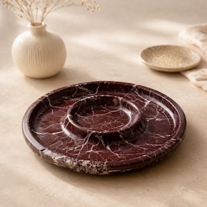 Puede incluir: Una fuente redonda de mármol burdeos con un cuenco central para salsas. La fuente tiene vetas blancas y un borde elevado. Un pequeño jarrón blanquecino con flores secas y un plato pequeño están en el fondo.