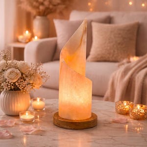 Puede incluir: Una lámpara de sal tallada y brillante, con un cálido brillo ámbar, sobre una base de madera. La lámpara está sobre una mesa de mármol con velas y pétalos de rosa. El fondo incluye un sofá y luces decorativas.