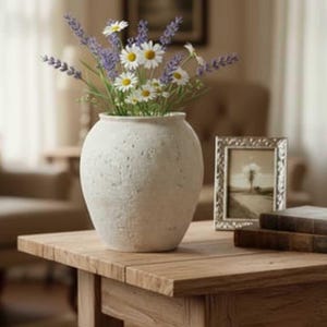 May include: A cream-coloured textured vase holding daisies and lavender sits on a wooden table. A framed picture and two books are also on the table. The room has a warm, inviting atmosphere.
