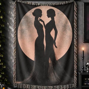 May include: A rectangular woven wall tapestry featuring a black silhouette of two figures standing face-to-face against a large, circular, pale peach-colored moon. The tapestry has a fringed border on the top and bottom edges and hangs from a black rod.