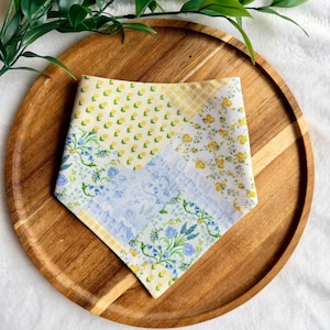May include: A triangular pet bandana featuring a patchwork design of various floral and gingham patterns in shades of yellow, blue, and white. The fabric displays small yellow flowers, blue botanical prints, and a yellow and white checkered border.