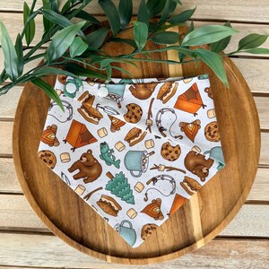 May include: A white triangular bandana featuring a repeating pattern of camping-themed illustrations including brown bears, orange tents, campfires, s'mores, chocolate chip cookies, fishing rods, pine trees, and blue mugs with two white snap buttons.