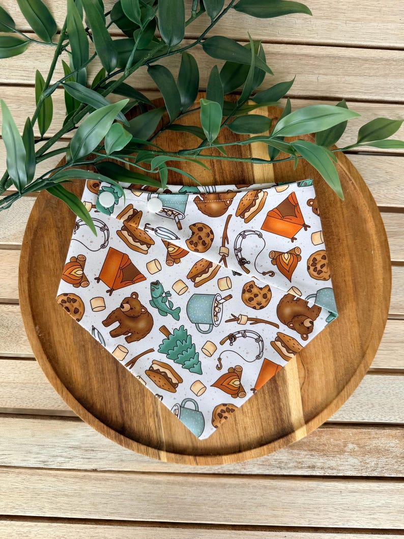 May include: A white triangular bandana featuring a repeating pattern of camping-themed illustrations including brown bears, orange tents, campfires, s'mores, chocolate chip cookies, fishing rods, pine trees, and blue mugs with two white snap buttons.