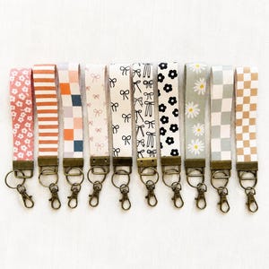 May include: Ten fabric wristlet keychains arranged in a row, each featuring a unique pattern including floral, striped, checkered, and bow designs. Each keychain is attached to a bronze-toned metal clasp and a circular split ring for keys.