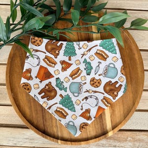 May include: A white triangular bandana featuring a repeating pattern of brown bears, green pine trees, orange tents, s'mores, chocolate chip cookies, blue mugs, fishing rods, and marshmallows scattered across the fabric surface.