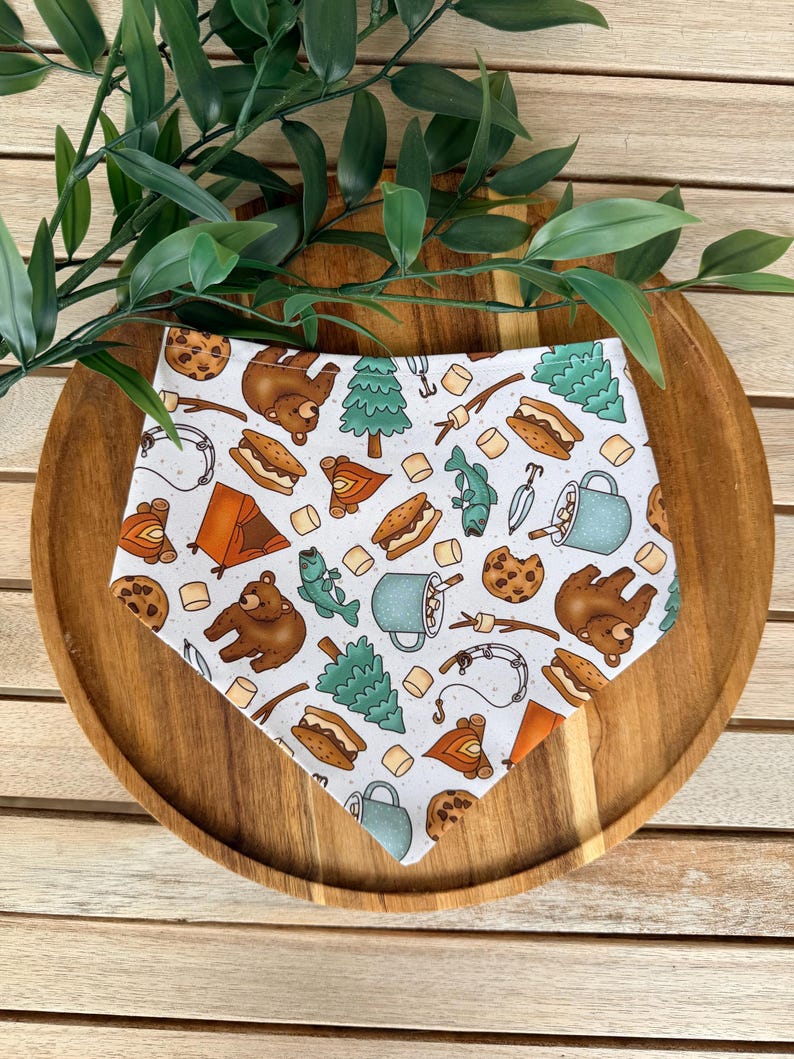 May include: A white triangular bandana featuring a repeating pattern of brown bears, green pine trees, orange tents, s'mores, chocolate chip cookies, blue mugs, fishing rods, and marshmallows scattered across the fabric surface.
