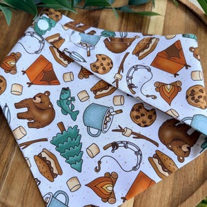 May include: A white triangular pet bandana featuring a repeating pattern of camping-themed illustrations including brown bears, orange tents, campfires, blue speckled mugs with marshmallows, chocolate chip cookies, fishing rods, and pine trees.