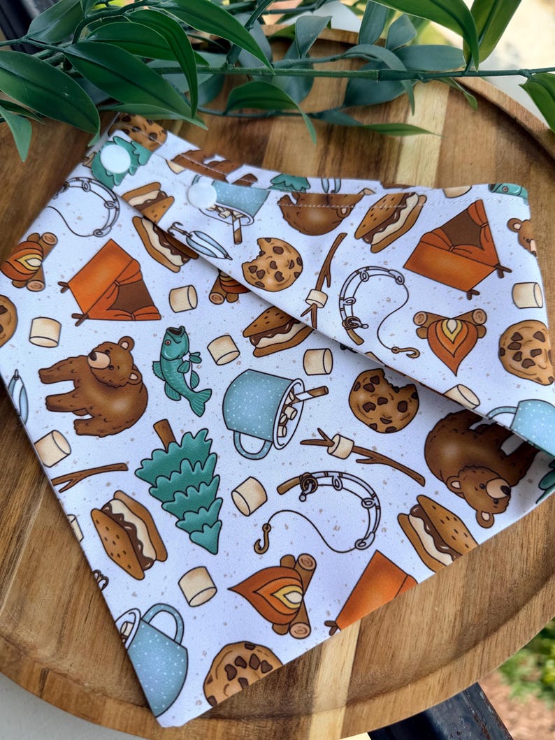 May include: A white triangular pet bandana featuring a repeating pattern of camping-themed illustrations including brown bears, orange tents, campfires, blue speckled mugs with marshmallows, chocolate chip cookies, fishing rods, and pine trees.