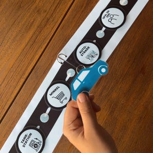 May include: A blue car-shaped keychain held near a black and white road map with circular icons. The map includes icons for a house, shop, and gas station. The background is a wooden surface.
