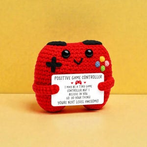 May include: A red crocheted game controller with black buttons and colorful buttons on the side. The controller holds a white sign that reads "POSITIVE GAME CONTROLLER" and "YOU'RE NEXT LEVEL AWESOME!"