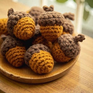 May include: A collection of handmade crochet acorns in shades of brown and golden yellow. The acorns are arranged on a wooden plate, showcasing their textured, detailed design. The craft project evokes a fall theme.
