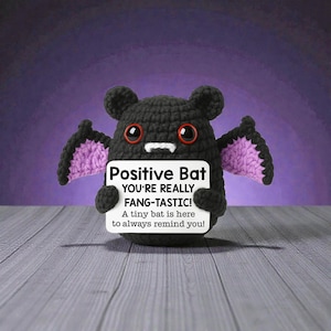 May include: A crocheted black bat with purple wing accents and red eyes holds a sign that reads "Positive Bat YOU'RE REALLY FANG-TASTIC! A tiny bat is here to always remind you!" The bat is on a wooden surface.
