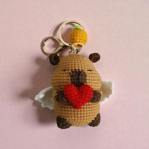 May include: A handmade crochet keychain featuring a brown bear holding a red heart. The bear has small white wings and a gold keychain ring. A small yellow pineapple charm hangs above the bear. The background is a solid pink.