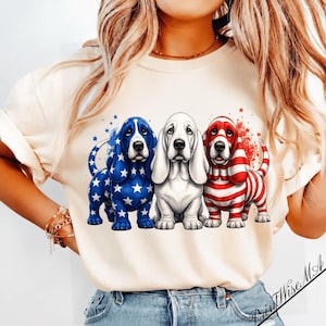Peut inclure: Un t-shirt couleur crème arborant un motif de trois chiens Basset Hound. Le chien de gauche est bleu avec des étoiles blanches, celui du centre est blanc uni, et celui de droite présente des rayures rouges et blanches. Ils ont de longues oreilles.