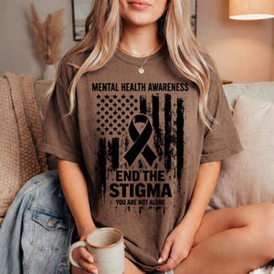 Peut inclure: T-shirt marron avec l'inscription "MENTAL HEALTH AWARENESS" au-dessus d'un ruban noir et d'un motif de drapeau américain. Le texte "END THE STIGMA" et "YOU ARE NOT ALONE" sont en dessous.