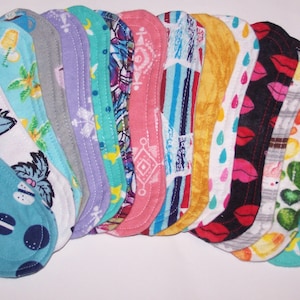 May include: A collection of reusable cloth menstrual pads in various patterns and colors. Designs include coffee cups, tropical leaves, geometric shapes, and lips. Each pad has a curved shape and is made of fabric.