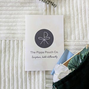 May include: A cream-coloured card with a dark grey circular logo and text reading "The Pippa Pouch Co. Scripture, held differently." A dark green pouch with a drawstring is partially visible, along with other items. The background is a white, textured fabric.