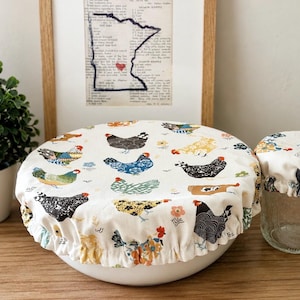 May include: Two fabric bowl covers with an off-white background featuring a repeating pattern of multi-colored chickens in shades of black, blue, yellow, and orange. The covers have elasticized edges and are fitted over a large bowl and a small glass jar.