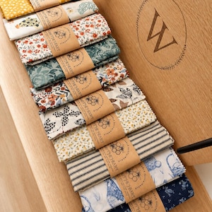 May include: A collection of ten folded fabric wraps with various patterns including floral, striped, and chicken motifs. Each wrap is secured with a brown paper band featuring a honeycomb logo and the text "THE HEART SUSTAINABLE".