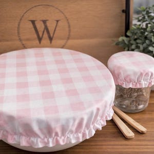 May include: Two round fabric bowl covers featuring a light pink and white gingham check pattern. The covers have elasticized, ruffled edges designed to fit snugly over containers. The set includes one large cover and one smaller matching cover.