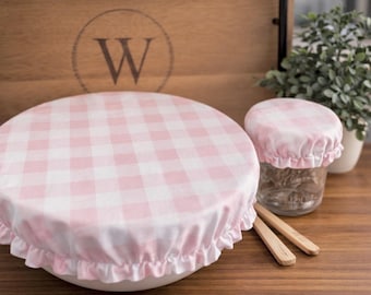May include: Two round fabric bowl covers featuring a light pink and white gingham check pattern. The covers have elasticized, ruffled edges designed to fit snugly over containers. The set includes one large cover and one smaller matching cover.