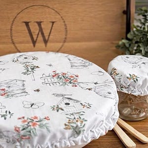 May include: White fabric bowl covers and jar covers with a bee and floral design. The covers have a ruffled edge and are placed on a wooden surface. A wooden box with a large "W" is in the background.