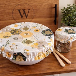 May include: A large and a small round fabric bowl cover featuring a repeating hexagonal pattern with yellow sunflowers, bees, and beehives on a white background. The covers have elasticized ruffled edges and are placed over a bowl and a glass jar.