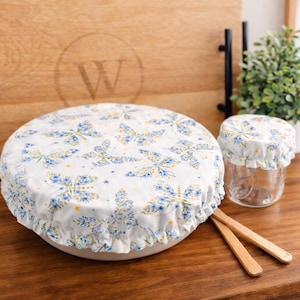 May include: White bowl cover and jar cover with a blue and yellow butterfly pattern. The bowl cover is ruffled around the edges. Two wooden utensils are next to the bowl. A wooden cutting board is in the background.