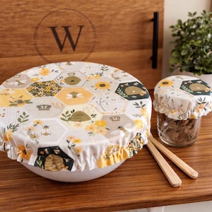 May include: A white bowl and a small jar are covered with reusable fabric bowl covers. The covers feature a hexagonal pattern with bees, sunflowers, and crowns. Wooden utensils are next to the jar. A wooden tray is in the background.