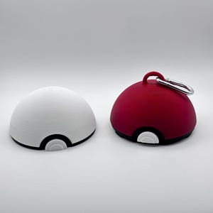 May include: Two spherical objects resembling a Pokeball. One is white with black and white accents, the other is red with black and white accents, and a silver carabiner. The objects are likely 3D printed.