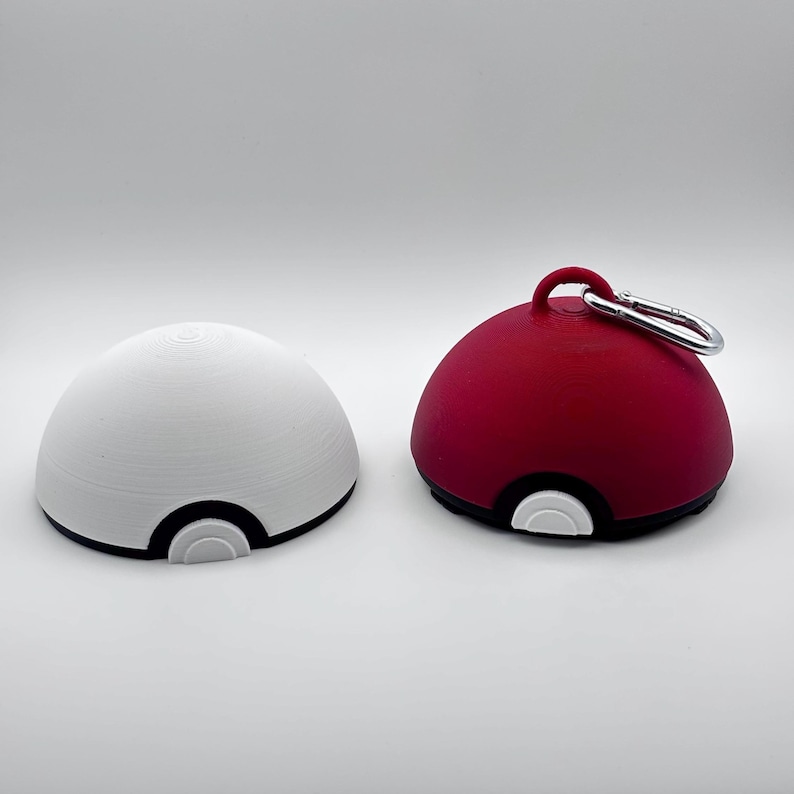 May include: Two spherical objects resembling a Pokeball. One is white with black and white accents, the other is red with black and white accents, and a silver carabiner. The objects are likely 3D printed.