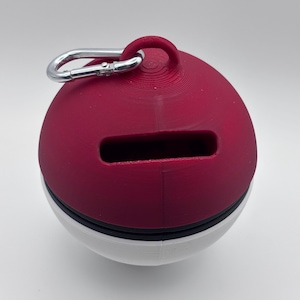 May include: A red and white sphere-shaped container with a rectangular slot and a silver carabiner clip. The top half is a deep red, the bottom half is white, and a black band separates the two. The container has a loop at the top.