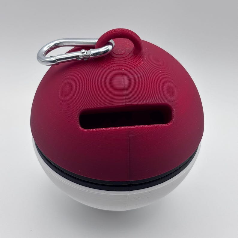 May include: A red and white sphere-shaped container with a rectangular slot and a silver carabiner clip. The top half is a deep red, the bottom half is white, and a black band separates the two. The container has a loop at the top.