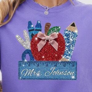 May include: A purple t-shirt featuring a school-themed graphic. The design includes a red glitter apple with a pink bow, blue crayons, a blue pencil, and scissors. A blue ruler with the text "Mrs. Johnson" is at the bottom.
