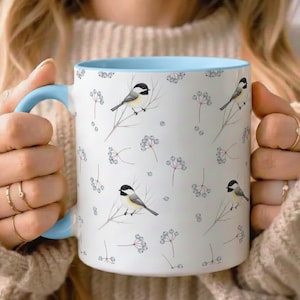 May include: White ceramic mug with a light blue interior and handle. The mug features a repeating pattern of small birds perched on branches with berries. The mug is held by a person wearing a beige sweater.