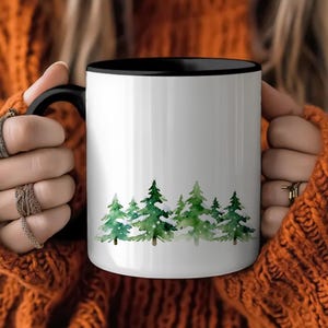 May include: White ceramic mug with a black interior and handle. The mug features a watercolor design of green pine trees at the bottom. The mug is held by a person wearing an orange sweater.