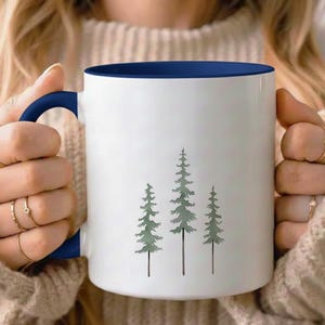 May include: White ceramic mug with a navy blue interior and handle. The mug features a minimalist design of three green pine trees. The mug is held by a person wearing a beige sweater.