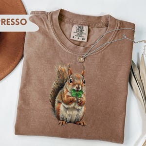 May include: Espresso-colored t-shirt featuring a detailed illustration of a squirrel holding a four-leaf clover. The shirt has a crew neck and short sleeves. A silver necklace with a pendant rests on the shirt. The shirt is made of a soft, comfortable material.
