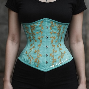 May include: A teal corset with gold floral embroidery. The corset has a structured design with silver-toned clasps down the front. The website address corsettly.com is printed on the bottom right of the corset.