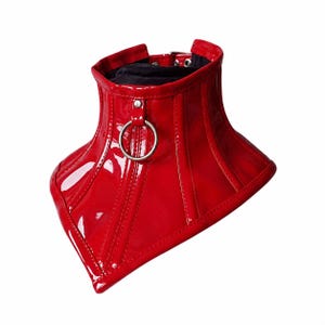 May include: A high-neck red patent leather choker featuring a glossy finish, vertical stitched paneling, and a central silver-toned metal O-ring. The interior is lined with black fabric, and the back includes a silver-toned metal buckle closure.