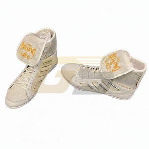 Puede incluir: Par de zapatillas de boxeo blanquecinas con detalles dorados. Las zapatillas de caña alta presentan el texto "Boxing Life" en dorado en la lengüeta. Las zapatillas tienen un diseño texturizado y están sobre un fondo blanco.