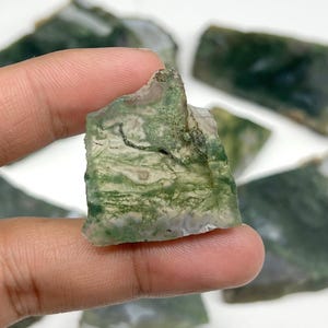 Puede incluir: Primer plano de piedras de ágata musgosa en bruto y natural. Las piedras tienen una mezcla de colores verdes y blancos con un patrón orgánico único. Los bordes ásperos y la textura natural son visibles. Varias piedras están dispersas.