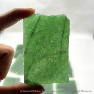 May include: A rectangular, translucent, green jade stone is held up against a white background. The stone has a rough, natural texture and varying shades of green. Other jade pieces are visible in the background. The text at the bottom of the image is excluded.
