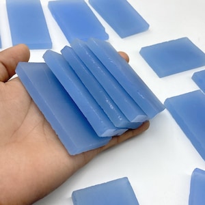 May include: A hand holding a stack of light blue rectangular bars, with several more scattered in the background. The bars appear to be made of a translucent material, possibly soap or resin.