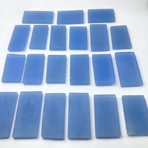 May include: A collection of light blue rectangular glass tiles are arranged in rows. Each tile has a slightly irregular shape and a smooth, translucent appearance. The color is a soft, muted blue. The tiles are displayed from an overhead perspective.